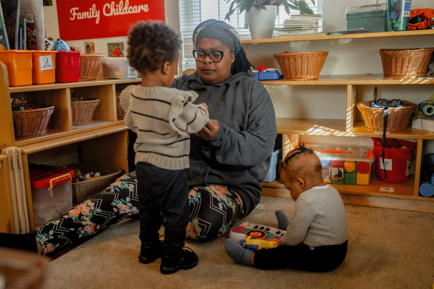 A woman plays with two young children in a vibrant classroom, surrounded by toys and cheerful decorations.