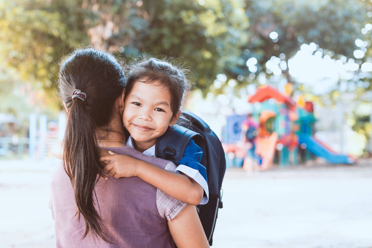 A young child similing over her mother‘s shoulder. There is a playground blurred out in the background.