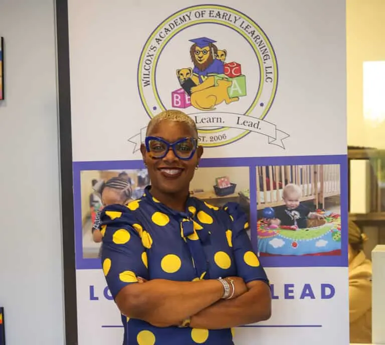 A woman in a blue and yellow polka dot dress poses confidently in front of a sign for a childcare academy.
