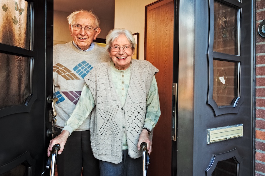 An elder man and woman smiling from their front door. The woman is using a walker.
