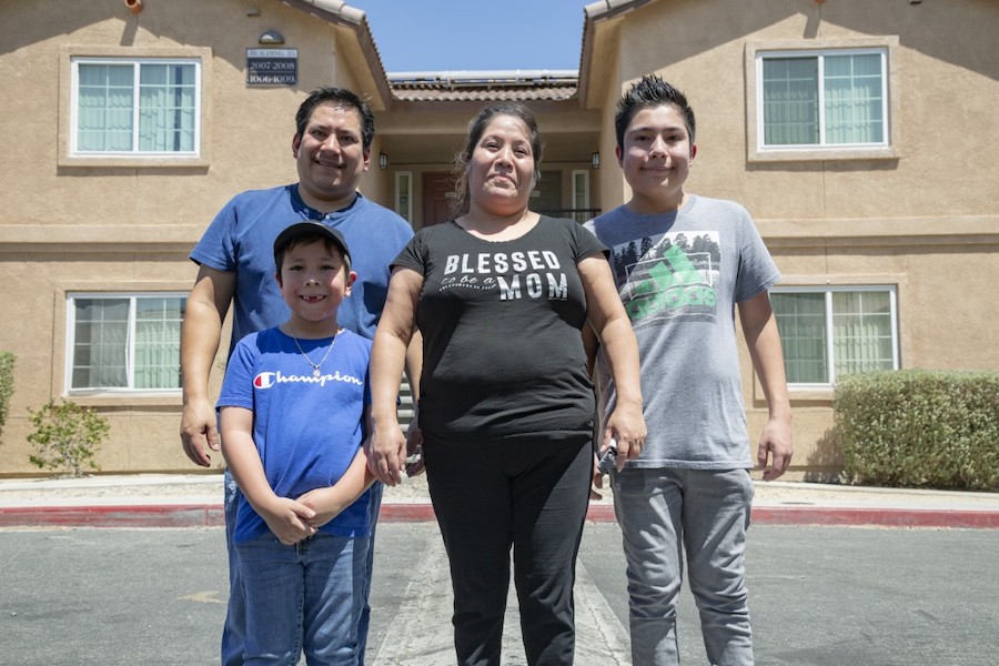 A Latino family (mother, father, and two young boys) smiling and standing together in front of their home.